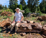 Man (Artist Robert C Paulson Jr [RC Paulson Jr]) sitting on a large log beside more falen wood in a wooded area with trees in the background. Stack of cut logs, one piece of which will become a beautiful bowl.  This is where the wood was gathered.