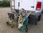 A green hiking backpack loaded with large pieces of driftwood roots, a black curved hand saw with a red handle, one trekking pole, and logs, positioned in front of the rear bumper of a white pickup truck parked on gravel.