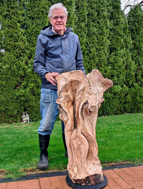 Artist Robert C Paulson Jr standing beside Freeform one-of-a-kind exotic sculpture still in its rough carving stage (but nearly finished), maple wood floor sculpture with asymmetrical twists and turns, grooves, see through sections, and unique cup-like shapes.  42.5 tall x 25 wide x 17''. Man holding a large wooden sculpture outdoors with greenery in the background