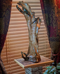 A polished 27 inch tall natural decorative Sitka spruce driftwood root sculpture with rich brown, beige, and golden orange patina tones, displayed on a wooden pedestal in front of window blinds and patterned curtains.