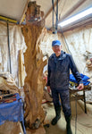 Artist and wood sculptor Bob Paulson standing in his shop beside his rich looking, one-of-a-kind, 7+ foot tall by 24" x 24'' natural spruce floor sculpture that is not yet completed.  When completed, it will emphasize the tree's natural form and beauty. It will have a curved and twisted shape, some live edge design and will be a combination of smooth and and a few slightly rough surfaces with beautiful rich colors and grain patterns.  