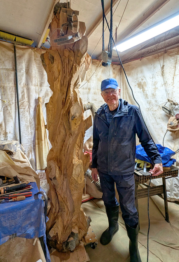 Artist and wood sculptor Bob Paulson standing in his shop beside his rich looking, one-of-a-kind, 7+ foot tall by 24" x 24'' natural spruce floor sculpture that is not yet completed.  When completed, it will emphasize the tree's natural form and beauty. It will have a curved and twisted shape, some live edge design and will be a combination of smooth and and a few slightly rough surfaces with beautiful rich colors and grain patterns.  