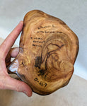 A hand holding the bottom of a sculpture that is a polished live-edge wood root with natural grain patterns, showing the artist's signature and handwritten information describing the sculpture (Pacific Ocean driftwood from Oregon, USA, Sitka spruce root), and the items SKU, 699-G.  