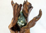 Close up view of the top of a 27 inch tall sculptural wooden piece resembling open hands or a shell, holding a green gray stone with white and gold barnacles, photographed against a plain off-white background.