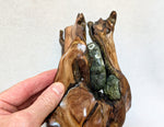 A hand holding close up view of the top of a 27 inch tall sculptural wooden piece resembling open hands or a shell, holding a green gray stone with white and gold barnacles, photographed against a plain off-white background.