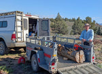 Artist Bob, Robert C Paulson, Jr., with the raw wood (still has its bark) and a chainsaw standing next to a truck with a trailer loaded with logs in a natural setting. 