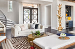 A 76 inch tall decorative tree sculpture standing in a modern home's living room. The artistic sculpture is twisting and short-branched, golden maroon, abstract and freeform. The room features a white sofa with neutral and black accent pillows, a geometric brown and beige rug, a round gold coffee table, and a white ottoman with a floral arrangement. A staircase is visible on the left, and dark wood flooring covers the room. 