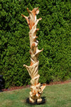 Potential left side view of a the completed 75.5" tall abstract branching indoor western juniper wood sculpture emphasizing the tree's original shapes.  It has beautiful rich grain patterns, natural unaltered golden tan and maroon colors, flowing flared lines with multiple grooves, intriguing hooked shapes.  One-of-a-kind 75.5" tall by 21" x 20'' floor sculpture.  It is smooth but has a few carefully conserved naturally rough surfaces. Seen here against a green conifer tree background.