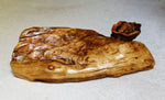 Bottom view of a 34 inch long natural burl wood decorative food tray charcuterie board with a polished finish, showing rich grain patterns and organic shapes, resting on a light gray textured surface.  Shows the artist's signature, wood species and sculpture's code number engraved. 