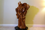 Rear or side 3 of a glossy, sculptured wooden decor piece shaped like a gnarled tree trunk, displayed on a round black rotating base (not included) in front of a beige wall. 
